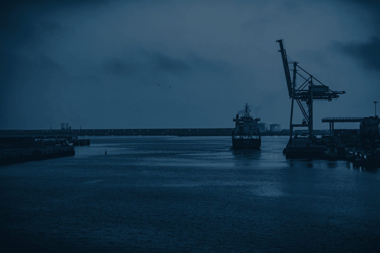 Cargo vessel leaving a commercial harbor at dusk, container crane on the quay